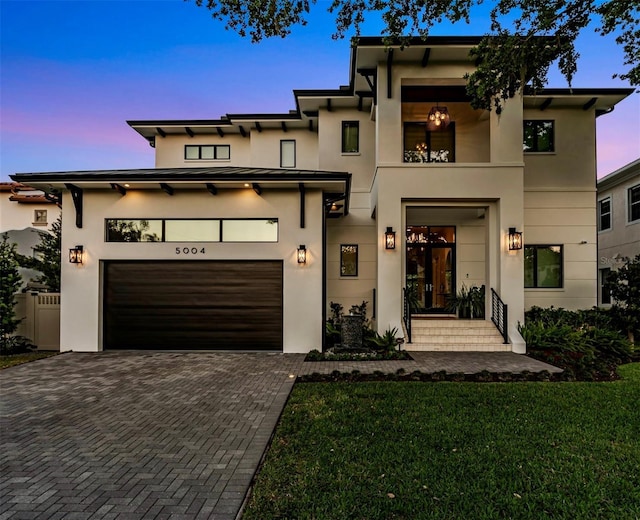 modern home featuring a front yard, an attached garage, a balcony, stucco siding, and decorative driveway