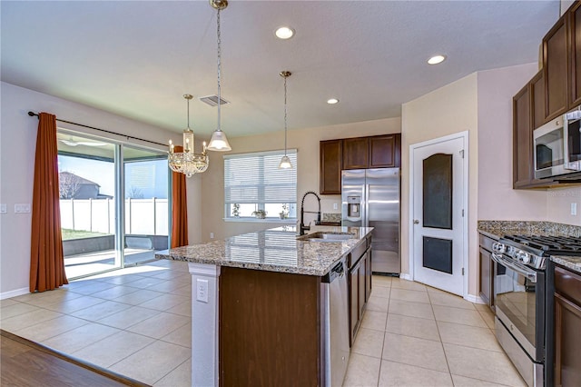 kitchen with sink, an island with sink, pendant lighting, stainless steel appliances, and light stone countertops