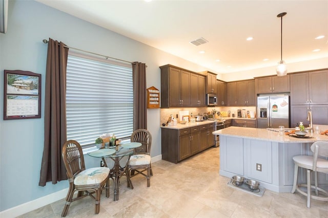 kitchen with pendant lighting, stainless steel appliances, light stone counters, a center island with sink, and decorative backsplash