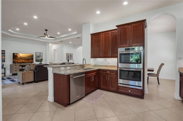 kitchen with sink, stainless steel appliances, a tray ceiling, light stone countertops, and kitchen peninsula