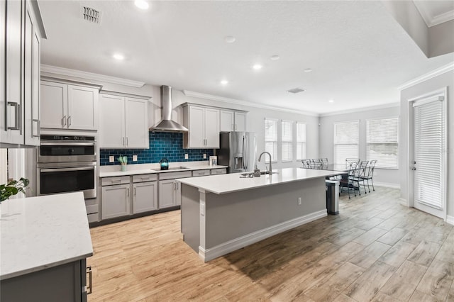 kitchen featuring stainless steel appliances, ornamental molding, wall chimney range hood, gray cabinets, and decorative backsplash