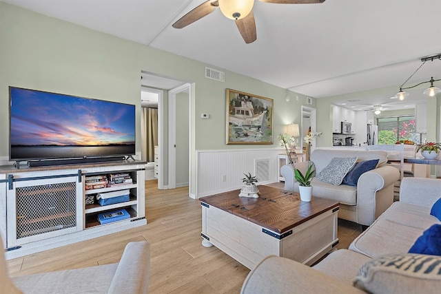 living area with light wood-type flooring, visible vents, a ceiling fan, and wainscoting