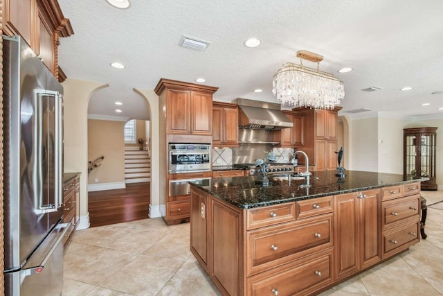 kitchen with appliances with stainless steel finishes, pendant lighting, ornamental molding, a center island with sink, and wall chimney exhaust hood