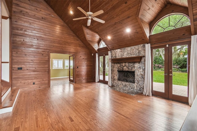 unfurnished living room featuring french doors, a stone fireplace, high vaulted ceiling, light wood-type flooring, and wooden walls