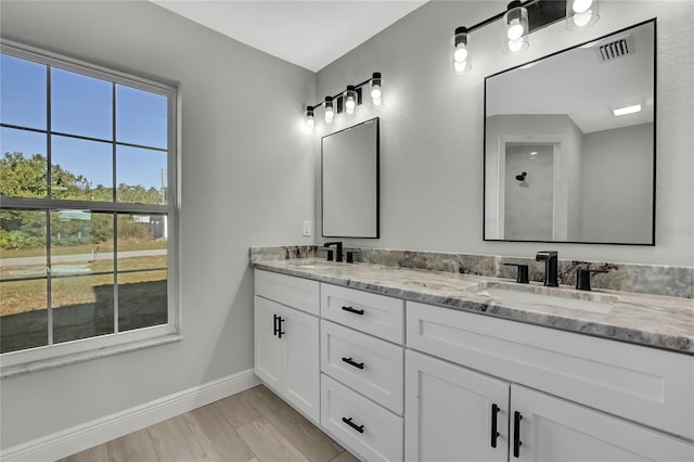 bathroom featuring a shower, double vanity, a sink, and baseboards