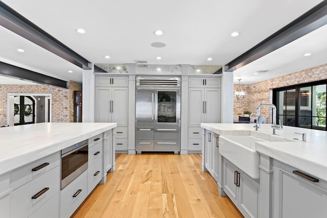 kitchen featuring beam ceiling, built in appliances, light stone countertops, brick wall, and light wood-type flooring