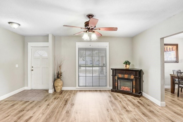 foyer entrance with light wood-type flooring, a glass covered fireplace, ceiling fan, and baseboards