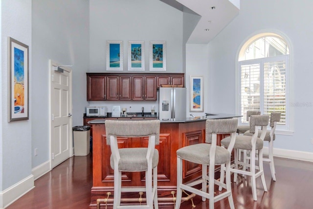 kitchen with a towering ceiling, white microwave, a breakfast bar, dark wood-style flooring, and stainless steel refrigerator with ice dispenser