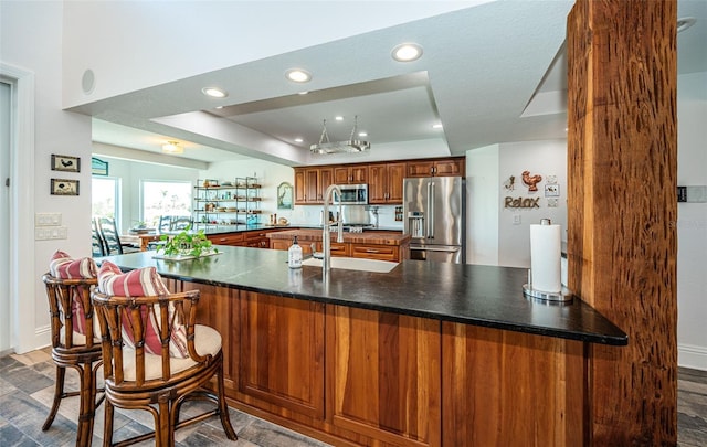 kitchen with dark countertops, a raised ceiling, appliances with stainless steel finishes, brown cabinetry, and a peninsula