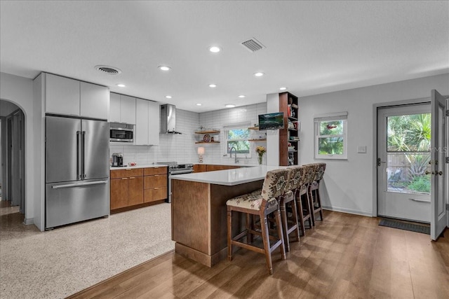 kitchen featuring wall chimney range hood, light hardwood / wood-style flooring, a breakfast bar area, stainless steel appliances, and decorative backsplash