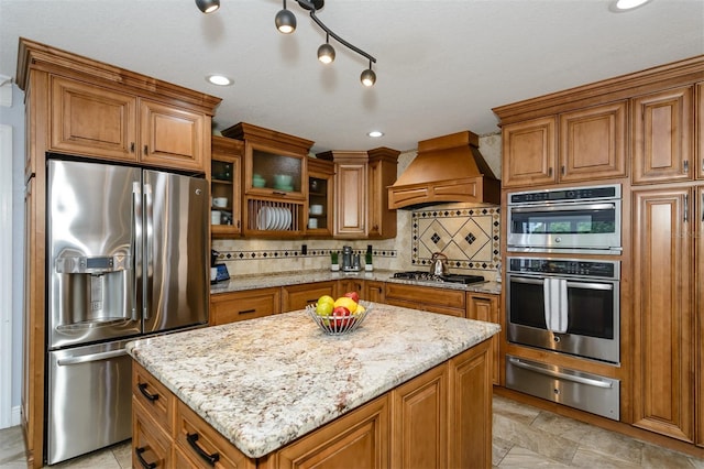 kitchen featuring premium range hood, appliances with stainless steel finishes, backsplash, a center island, and light stone counters