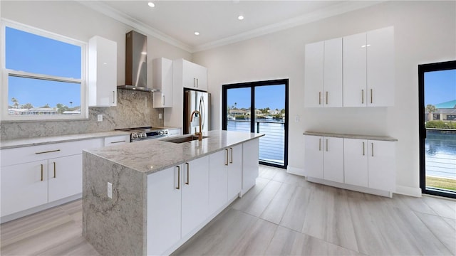 kitchen with wall chimney exhaust hood, sink, a water view, an island with sink, and white cabinets