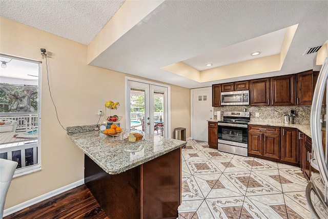 kitchen featuring appliances with stainless steel finishes, a tray ceiling, light stone countertops, kitchen peninsula, and french doors