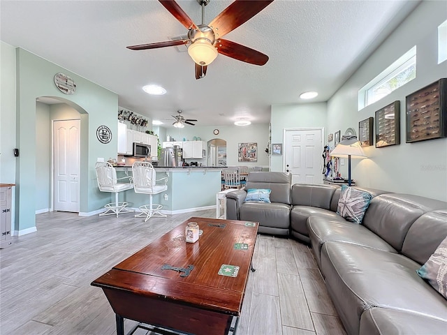 living room with a textured ceiling, arched walkways, light wood finished floors, baseboards, and ceiling fan