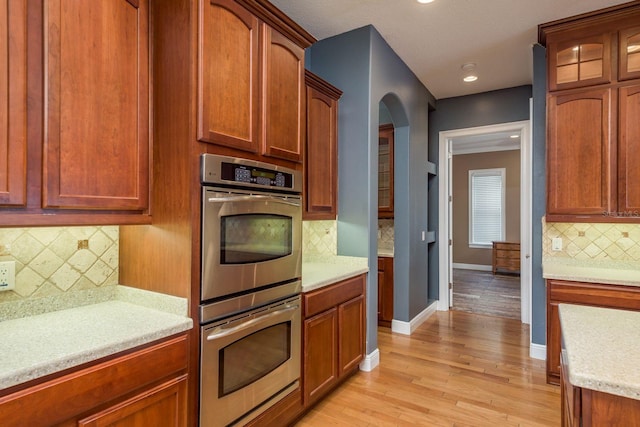 kitchen featuring baseboards, arched walkways, light wood-style flooring, stainless steel double oven, and backsplash