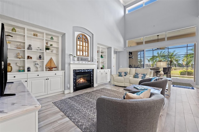 living room featuring built in shelves, a glass covered fireplace, light wood-style flooring, and a high ceiling