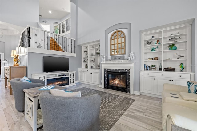 living area featuring light wood finished floors, built in features, a glass covered fireplace, a towering ceiling, and stairway