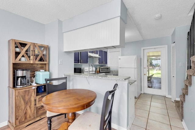 kitchen with sink, white refrigerator, kitchen peninsula, light stone countertops, and blue cabinetry