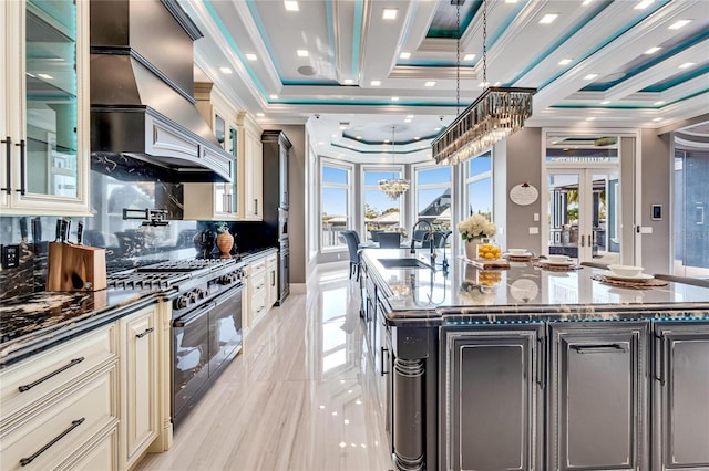 kitchen with range with two ovens, an island with sink, ornamental molding, dark stone countertops, and cream cabinetry