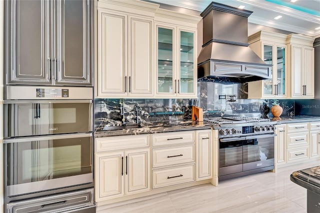 kitchen with stainless steel appliances, dark stone counters, cream cabinetry, a warming drawer, and custom range hood