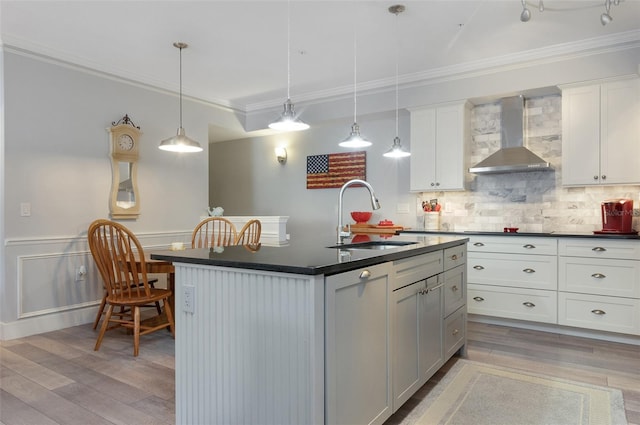 kitchen with pendant lighting, dark countertops, a sink, wall chimney range hood, and an island with sink