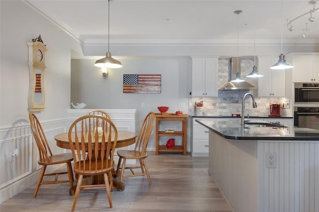 kitchen with a sink, white cabinetry, wall chimney range hood, dark countertops, and pendant lighting