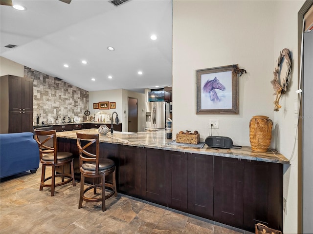 kitchen with vaulted ceiling, light stone counters, stainless steel refrigerator with ice dispenser, a kitchen breakfast bar, and kitchen peninsula