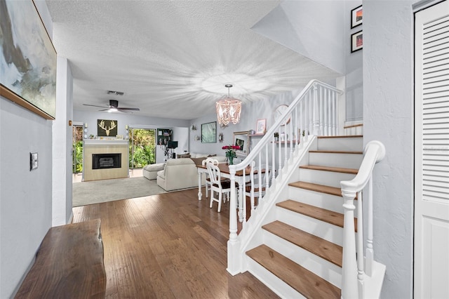 stairway featuring visible vents, wood-type flooring, a textured ceiling, a fireplace, and ceiling fan with notable chandelier