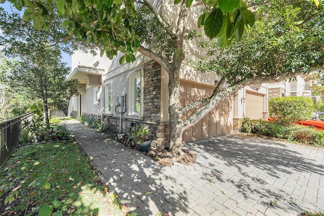 view of side of property featuring decorative driveway, stucco siding, an attached garage, fence, and stone siding