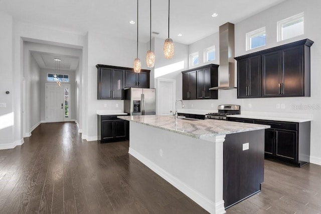 kitchen with dark wood-style flooring, appliances with stainless steel finishes, a sink, an island with sink, and wall chimney exhaust hood