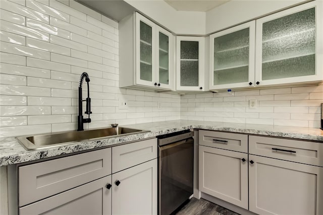 kitchen with tasteful backsplash, glass insert cabinets, light stone countertops, stainless steel dishwasher, and a sink