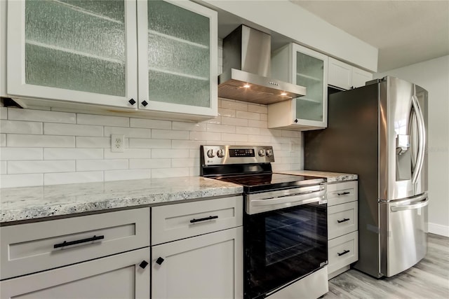 kitchen featuring light stone counters, light wood-style flooring, stainless steel appliances, backsplash, and wall chimney exhaust hood