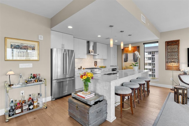 kitchen with white cabinets, hanging light fixtures, stainless steel appliances, a center island with sink, and wall chimney exhaust hood