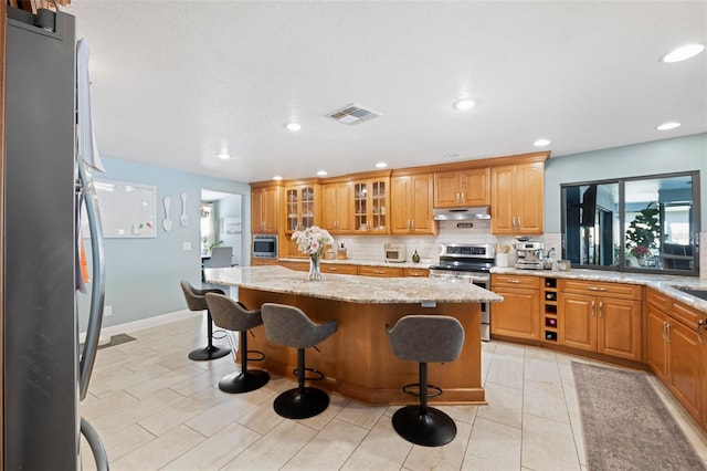 kitchen with appliances with stainless steel finishes, tasteful backsplash, an island with sink, a breakfast bar area, and light stone countertops