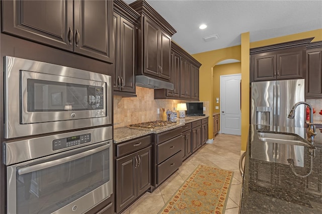 kitchen with arched walkways, stone counters, under cabinet range hood, appliances with stainless steel finishes, and tasteful backsplash