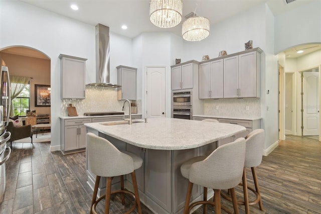 kitchen featuring arched walkways, a center island with sink, gray cabinets, double oven, and wall chimney exhaust hood