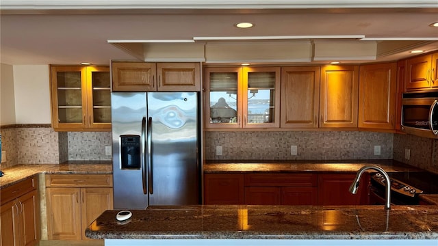 kitchen featuring stainless steel appliances, dark stone counters, brown cabinets, and glass insert cabinets