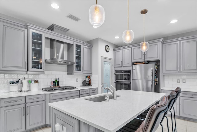 kitchen with glass insert cabinets, stainless steel appliances, a sink, wall chimney range hood, and a center island with sink