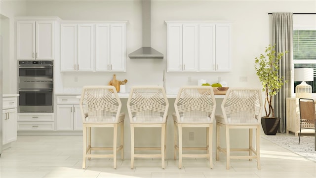 kitchen featuring stainless steel double oven, wall chimney exhaust hood, white cabinetry, and a breakfast bar area