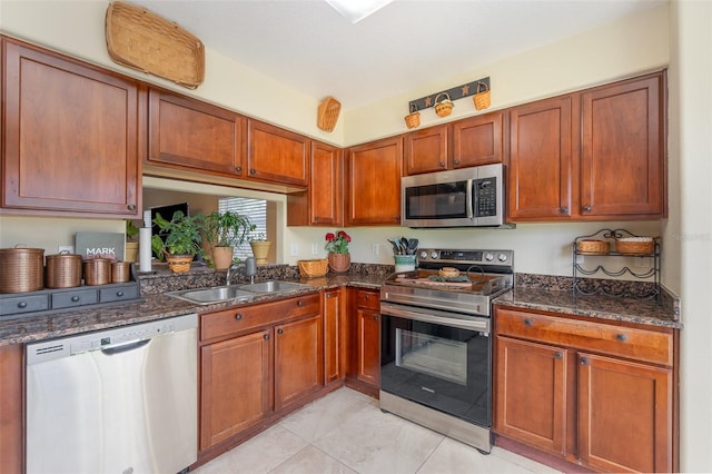 kitchen featuring brown cabinetry, appliances with stainless steel finishes, dark stone countertops, a sink, and light tile patterned flooring