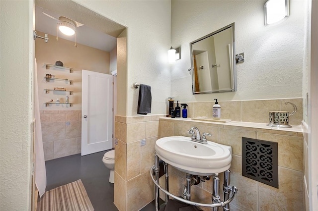 bathroom with tile walls, a textured wall, toilet, wainscoting, and a sink