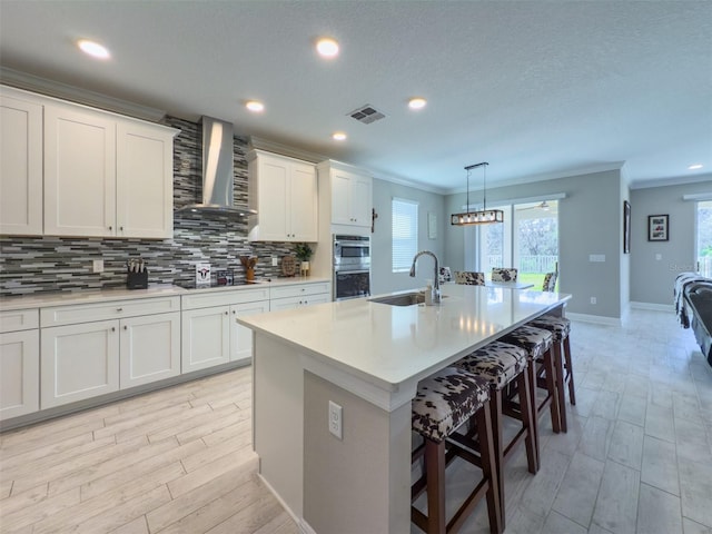 kitchen with light countertops, a kitchen island with sink, white cabinets, a sink, and wall chimney exhaust hood