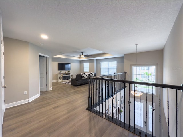 hall featuring baseboards, a raised ceiling, wood finished floors, a textured ceiling, and an upstairs landing