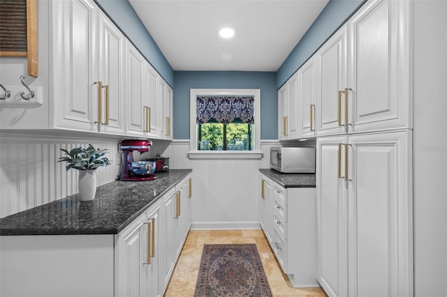 kitchen with dark stone counters, a wainscoted wall, stainless steel microwave, and white cabinetry