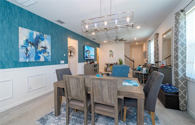 carpeted dining space with visible vents, stairway, wainscoting, ceiling fan, and wallpapered walls