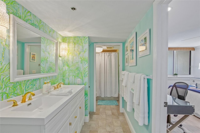 full bathroom with baseboards, double vanity, a sink, and wallpapered walls