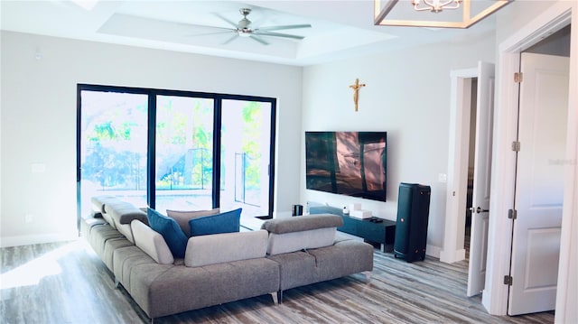 living room featuring ceiling fan with notable chandelier, a tray ceiling, wood finished floors, and baseboards