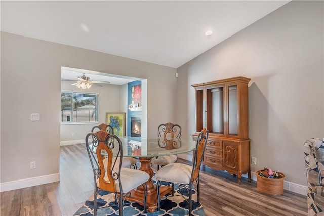 dining space with lofted ceiling, a glass covered fireplace, ceiling fan, wood finished floors, and baseboards