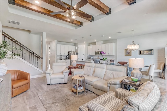 living room with ceiling fan with notable chandelier, visible vents, stairs, light wood-type flooring, and beamed ceiling