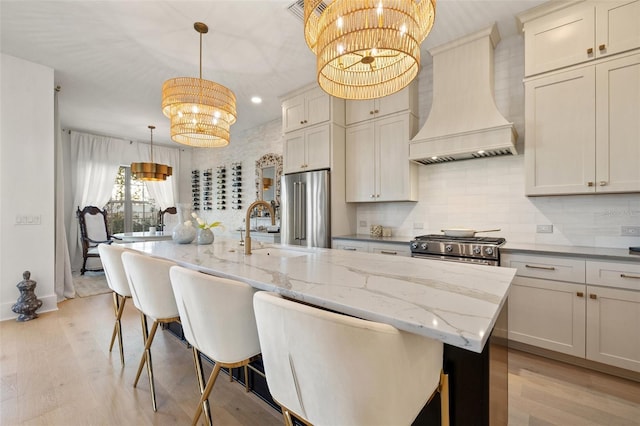 kitchen featuring premium appliances, a sink, hanging light fixtures, an island with sink, and custom range hood